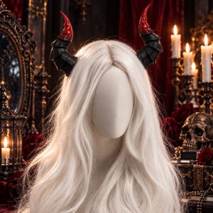 May include: A pair of segmented, curved horns featuring a matte black base that transitions into a glossy, vibrant red tip. The horns are attached to a headband and are displayed on a mannequin head with long, straight, platinum blonde hair.