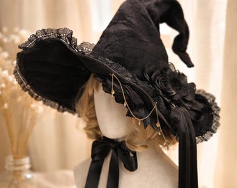 May include: Black witch hat with a wide brim, adorned with lace trim, a large fabric rose, and dangling gold-colored chains. The hat has a tall, pointed crown and black satin ribbons. A black bow is at the base of the hat.