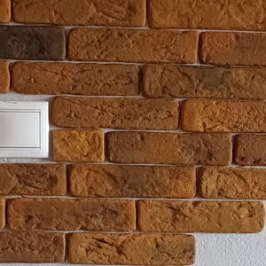 Puede incluir: Primer plano de una pared de ladrillos con diferentes tonos de marrón y beige. Un interruptor de luz blanco está centrado en la imagen. Los ladrillos tienen una superficie texturizada, creando una apariencia rústica.