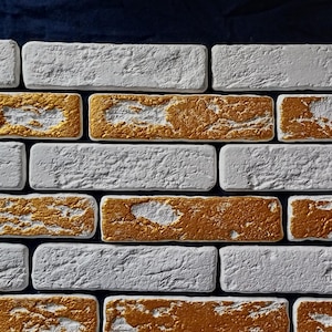 Puede incluir: Primer plano de un patrón de pared de ladrillos. El diseño presenta ladrillos rectangulares en filas alternas de blanco y dorado. Los ladrillos dorados tienen una apariencia texturizada y metálica, mientras que los ladrillos blancos tienen una textura rugosa similar a la piedra.