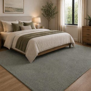 May include: A bedroom scene featuring a bed with a beige headboard, white bedding, and olive green accents. A large, rectangular, gray rug covers the floor. The room is decorated with wooden furniture, lamps, and a framed landscape painting.