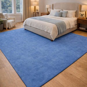May include: A large, rectangular, solid blue rug dominates the foreground of a bedroom scene. The rug's texture is visible, and it contrasts with the light-colored wooden floor. A bed with white bedding and blue accents is in the background.