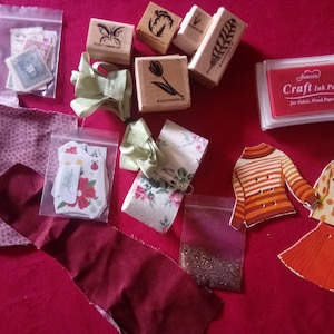 Puede incluir: Un conjunto de suministros para manualidades que incluye cuatro sellos de madera con diseños de mariposa, flor y pluma, una almohadilla de tinta roja con la etiqueta Craft Ink Pad, retazos de tela, cintas, recortes de papel y purpurina.