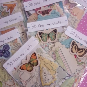 May include: Multiple clear plastic bags containing 30 pieces of map cutouts each. Each bag is labeled with the text 30 pieces Map cutouts and features a decorative butterfly or bird cutout placed on the front of the assorted paper shapes.