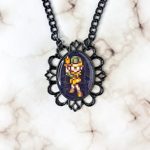 Puede incluir: Un collar de cadena negra con un colgante de metal negro que presenta un personaje pixelado con un sombrero verde y un atuendo amarillo. El colgante está rodeado por un marco decorativo de metal negro.