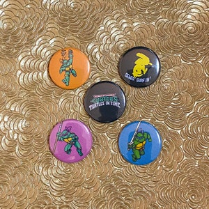May include: Four round pinback buttons featuring Teenage Mutant Ninja Turtles characters. The buttons are orange, black, purple, and blue. The orange button features a green turtle with a red bandana. The black button features the Teenage Mutant Ninja Turtles logo with the text "TURTLES IN TIME". The purple button features a green turtle with a purple bandana. The blue button features a green turtle with a blue bandana.