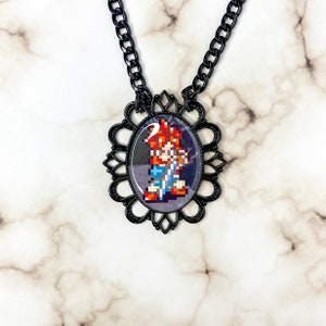Puede incluir: Collar negro con un camafeo que presenta un personaje pixelado con cabello rojo y una camisa azul. El camafeo está rodeado por un diseño de filigrana negro.