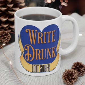 Puede incluir: Taza de cerámica blanca con el texto "WRITE DRUNK EDIT SOBER" en naranja y azul. La taza tiene forma de corazón azul con una pancarta amarilla. La taza está llena de un líquido oscuro y está sobre una superficie gris claro.