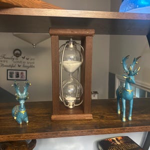 May include: An hourglass with white sand inside a clear glass bulb, encased in a dark brown wooden frame. Two teal deer figurines with gold accents flank the hourglass on a dark brown shelf. A book is visible on the shelf.