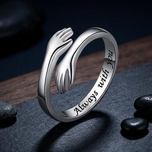 May include: A silver-toned open ring featuring two hands meeting in a hug design. The outer band is engraved with the black text Always with you. The ring has a polished metallic finish and a continuous band that wraps around the finger.