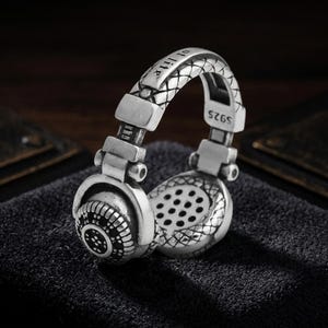 May include: A silver-colored headphone ring with intricate detailing. The ring features a textured headband with the inscription "of life". The earcups have a detailed design with small holes and a textured pattern. The ring is marked with "S925".