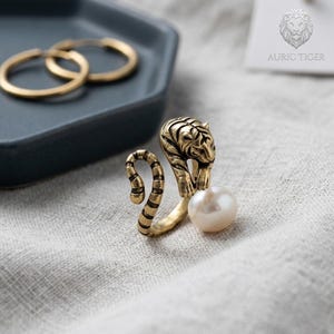 May include: A gold-colored ring featuring a tiger design with a pearl. The ring has a detailed tiger head and a curved tail. The pearl is white and round. Gold hoop earrings are in the background.