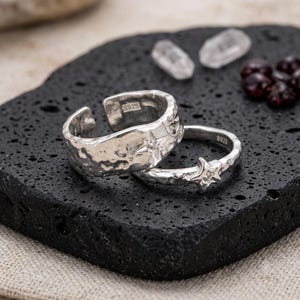 May include: Two silver-colored rings with an open-band design and a hammered texture. The wider ring features a star motif with a small embedded stone, while the thinner ring has a prominent star shape with a central stone. Both rings are stamped S925.