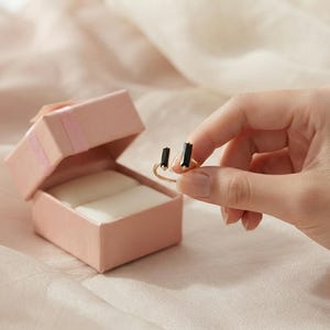 May include: A gold-toned open ring featuring two rectangular black stones at the ends, positioned next to an open, light pink square jewelry box with a white interior cushion, designed to hold small accessories securely for display or storage.
