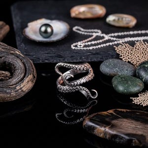 May include: A silver octopus tentacle ring is the focal point, with a black pearl in a shell and a silver necklace. The ring's design is detailed, and the scene includes stones and coral, all on a black surface.