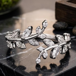 May include: A silver-toned bypass ring featuring two overlapping leaf-shaped branches. Each branch is adorned with multiple small, clear, round-cut and marquise-cut stones that mimic the appearance of sparkling foliage along the metal stems.