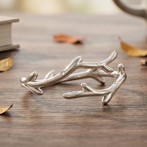 May include: Silver-colored, twig-shaped bracelet with small, clear embellishments. The bracelet is open and rests on a wooden surface. The design resembles a natural branch, with multiple points extending outwards. The bracelet is a decorative accessory.