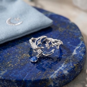 May include: A silver ring with a celestial design featuring a crescent moon, stars, and a dangling blue gem. The ring has a blue enamel inlay and is set on a blue surface. A small blue pouch with a crescent moon and star is in the background.