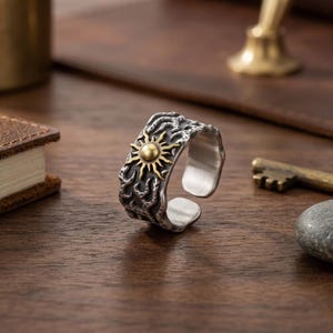 May include: A silver-toned adjustable ring with a textured band and a gold-colored sun design. The sun has a central circle with radiating lines. The ring is displayed on a wooden surface.
