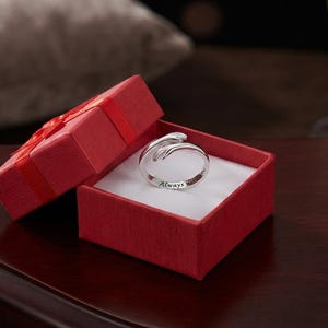 May include: A silver-toned metal ring shaped like two hands embracing, featuring the engraved text Always on the inner band, presented inside a square red gift box with a matching red ribbon on the lid and a white interior lining.