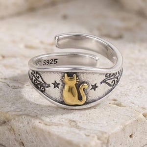 May include: A silver-tone adjustable ring with a gold-colored cat design. The ring features a decorative band with stars and swirls. The inscription "S925" is visible on the ring's surface.