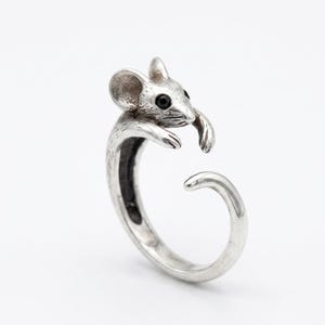 May include: A silver-colored ring in the shape of a mouse. The mouse has detailed features, including small black eyes and a textured surface. The ring wraps around the finger, with the mouse's head and paws forming the top.