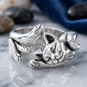 May include: A silver-toned metal ring shaped like a sleeping cat. The band features the cat's curled tail on one side and its head with tucked paws on the other, creating an open-ended design with detailed engravings of fur and facial features.