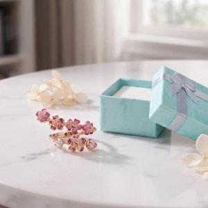 May include: A rose gold ring featuring a spiral design adorned with pink and white enamel flower motifs and small clear stones, positioned next to an open light blue square gift box with a matching ribbon bow and white dried flower petals.