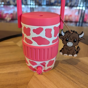 May include: A pink and white cow-print beverage cooler with a top handle, a pink lid, and a small spigot at the base. A brown highland cow charm hangs from a beaded chain attached to the handle, featuring white horns and a detailed cartoon face.