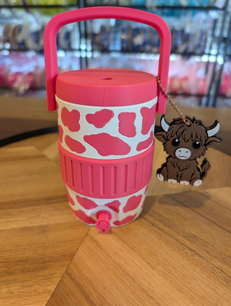 May include: A pink and white cow-print beverage cooler with a top handle, a pink lid, and a small spigot at the base. A brown highland cow charm hangs from a beaded chain attached to the handle, featuring white horns and a detailed cartoon face.