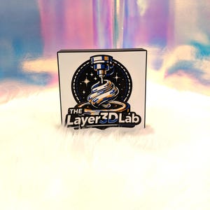 Op de afbeelding: Een vierkant bord met zwarte rand, met een 3D-printerafbeelding in blauw, wit en oranje tegen een sterrenachtergrond. De tekst "The Layer3D Lab" staat onder het centrale ontwerp.