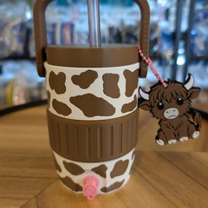 May include: A brown and white cow-print tumbler featuring a brown handle, a clear straw, and a pink spigot at the base. A small, brown, cartoon-style Highland cow charm hangs from a pink beaded chain attached to the side of the lid.