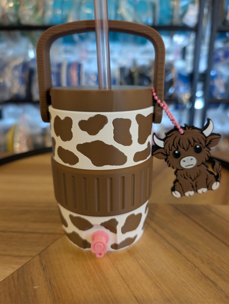 May include: A brown and white cow-print tumbler featuring a brown handle, a clear straw, and a pink spigot at the base. A small, brown, cartoon-style Highland cow charm hangs from a pink beaded chain attached to the side of the lid.