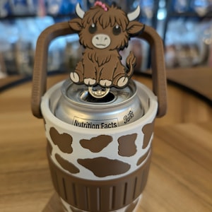 May include: A white and brown cow-print can cooler featuring a brown handle and a pink circular accent at the base. A brown highland cow figure with white horns and a small pink flower on its head is attached to the top of the handle.