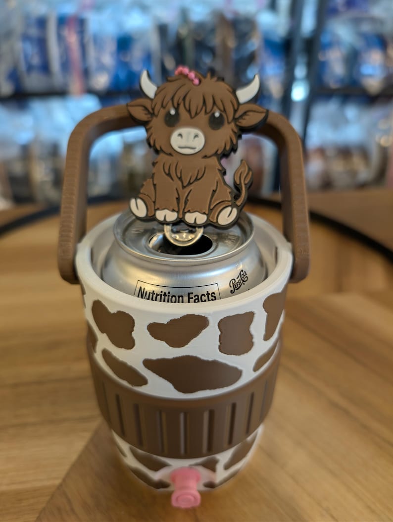 May include: A white and brown cow-print can cooler featuring a brown handle and a pink circular accent at the base. A brown highland cow figure with white horns and a small pink flower on its head is attached to the top of the handle.