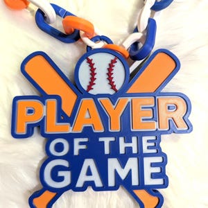 May include: A blue, white, and orange baseball-themed necklace. The pendant reads "PLAYER OF THE GAME" in orange and white lettering, with a baseball and crossed bats design. The chain links are blue, white, and orange.