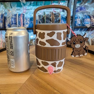 May include: A white and brown cow-print beverage cooler with a brown handle and a pink spigot. A brown highland cow charm hangs from the handle by a pink beaded chain. A silver aluminum beverage can is positioned next to the cooler for scale.