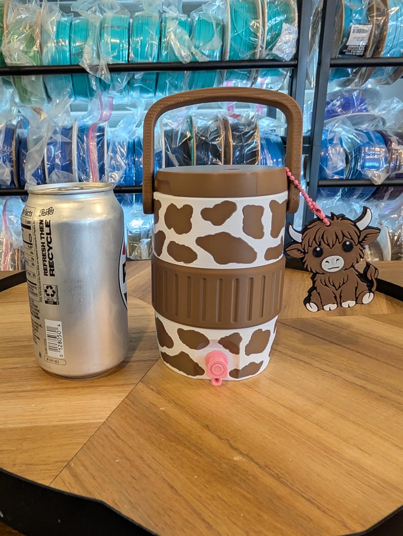 May include: A white and brown cow-print beverage cooler with a brown handle and a pink spigot. A brown highland cow charm hangs from the handle by a pink beaded chain. A silver aluminum beverage can is positioned next to the cooler for scale.