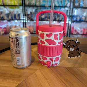 May include: A pink and white cow-print insulated beverage cooler with a handle, a clear straw, and a small spigot at the base. A brown highland cow charm hangs from the handle. A standard silver aluminum soda can is placed next to the cooler.
