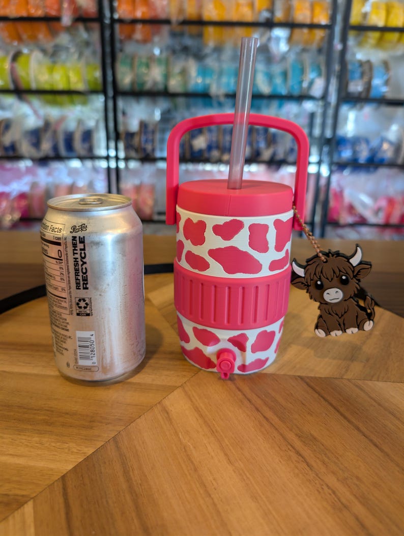 May include: A pink and white cow-print insulated beverage cooler with a handle, a clear straw, and a small spigot at the base. A brown highland cow charm hangs from the handle. A standard silver aluminum soda can is placed next to the cooler.