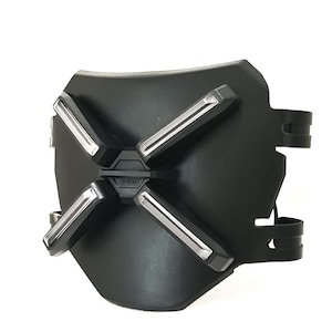 Può includere: Un dispositivo da polso nero dall'aspetto futuristico con quattro accenti argentati a forma di X. Il dispositivo ha una forma curva, a scudo, e include cinturini regolabili. Il design suggerisce un tema tecnologico o fantascientifico.