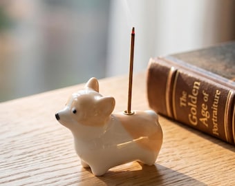 May include: A ceramic incense holder shaped like a Corgi dog with white and tan coloring. The figurine features a small hole on its back where a thin, brown incense stick is inserted, emitting a visible trail of smoke into the air above it.