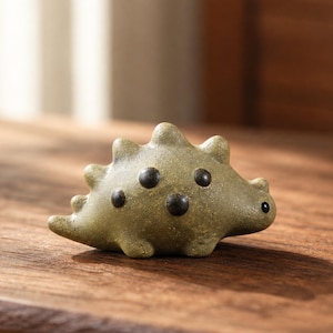 May include: A small, olive-green ceramic figurine shaped like a stylised dinosaur. The creature features a textured surface, a series of rounded dorsal spikes along its back, and three distinct dark brown circular spots on its side.