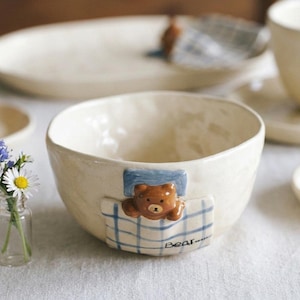 Pode incluir: Tigela de cerâmica creme com um desenho de urso a dormir. O urso castanho está a descansar numa almofada azul e num cobertor xadrez. A palavra "Bear" está escrita no cobertor. Outras peças de cerâmica e flores no fundo.