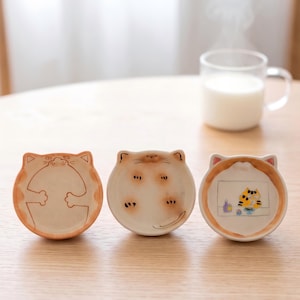 Op de afbeelding: Drie kleine, ronde keramische schaaltjes in de vorm van kattenkoppen. Het linker schaaltje toont een oranje omrande kat met de pootjes tegen elkaar. Het middelste schaaltje toont een lichtbruine kat met pootjes en staart. Het rechter schaaltje toont een oranje kat.