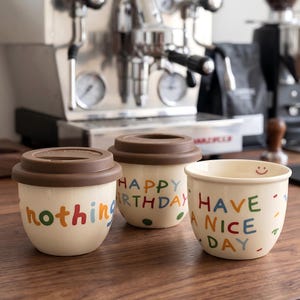 Op de afbeelding: Drie crèmekleurige keramische koffiekopjes met bruine deksels en kleurrijke tekst. Eén kopje zegt "nothing", een ander "Happy Birthday" en de derde "Have a Nice Day" met een smiley. De kopjes staan op een houten oppervlak.