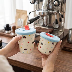 Peut inclure: Deux tasses en céramique crème avec des couvercles bleu clair et des accents rouges. Les tasses sont décorées de points colorés et d'un visage de dessin animé. Une tasse a une paille transparente. Les tasses sont sur une surface en bois près d'une machine à café.