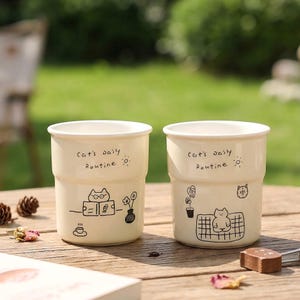 Puede incluir: Dos tazas de cerámica color crema con dibujos lineales negros de gatos y el texto "Cat's daily Routine". Una taza muestra un gato leyendo, la otra un gato relajándose. Las tazas están sobre una mesa de madera.