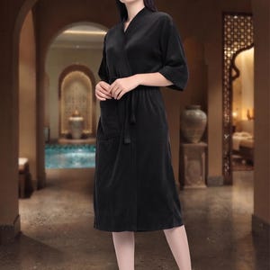 May include: A solid black robe featuring a V-neckline, three-quarter length sleeves, and a matching fabric tie belt at the waist. The garment has a knee-length hem and a patch pocket on the front right side, constructed from a soft, textured fabric.