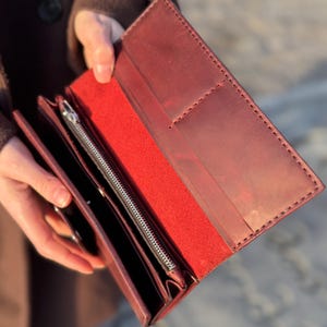 May include: Burgundy leather wallet with a red interior, held open to display its contents. The wallet features a silver zip and multiple card slots. The edges are stitched. The wallet is made of leather.